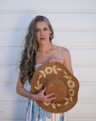 Model wearing 5.5" Brim Gigi Cinnamon and Natural Floral Embroidered Crochet Straw Sun Hats handmade crochet natural raffia palm fiber in a solid hue of cinnamon dark brown making a concentric pattern from the center top of the flattened crown spiraling out to the edge of the brim with matching adjustable raffia braid hat band and natural straw color floral pattern with leaves embroidered on the brim - Shebobo