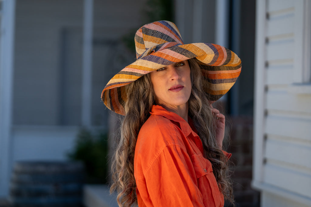 Model wearing 7" Wide Brim Candyland Multicolor Stripes Packable Straw Sun Hat handmade loomed raffia in bands of natural straw color, black, saffron yellow/orange, light pink, coral orange/red, and dusk navy/dark/blue stripes create a swirl pattern - Shebobo