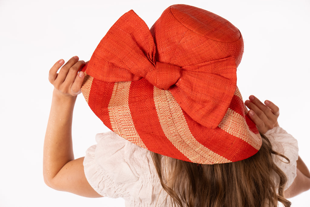 Model wearing 5" Wide Brim Coral Two Tone Wide Stripe Straw Sun Hats with Big Bow handmade loomed raffia palm fiber in a solid hue of Coral orange/red on the crown and matching oversized big bow embellishment hat band, with two tone even width wide stripe pattern of coral and natural straw color that creates a swirl patterned brim - Shebobo