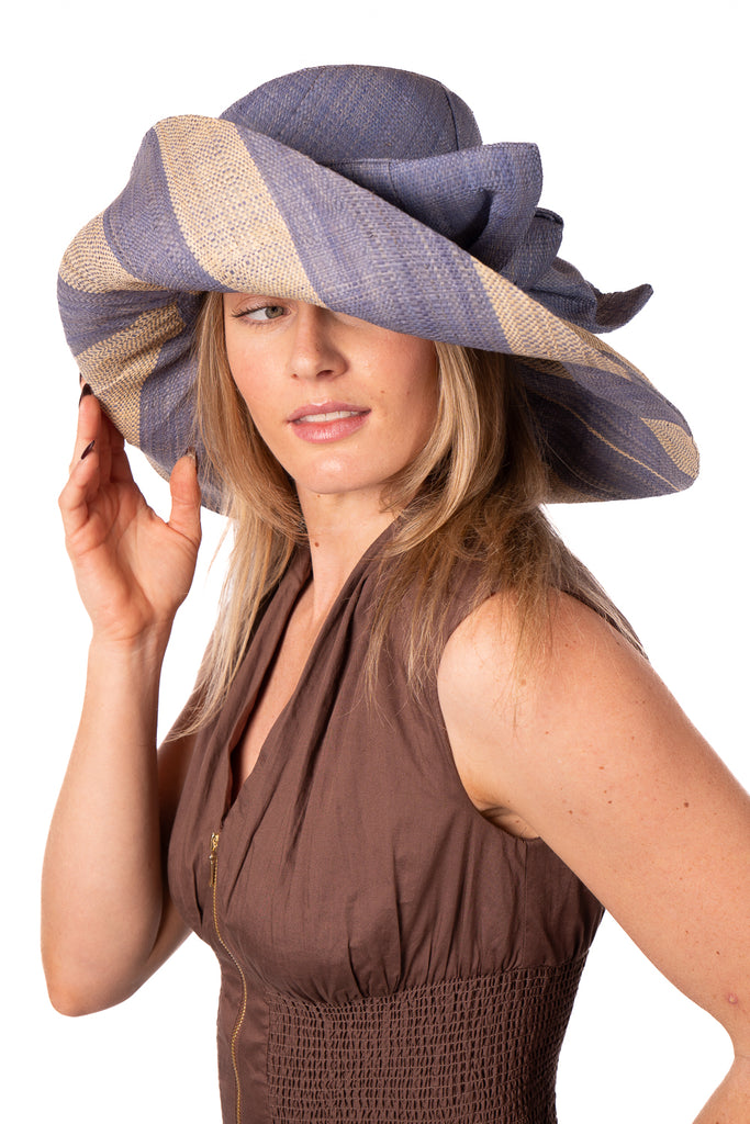 Model wearing 7" Wide Brim Denim Two Tone Wide Stripe Straw Sun Hats with Big Bow handmade loomed raffia palm fiber in a solid hue of Denim retro/light/blue on the crown and matching oversized big bow embellishment hat band, with two tone even width wide stripe pattern of denim and natural straw color that creates a swirl patterned brim - Shebobo