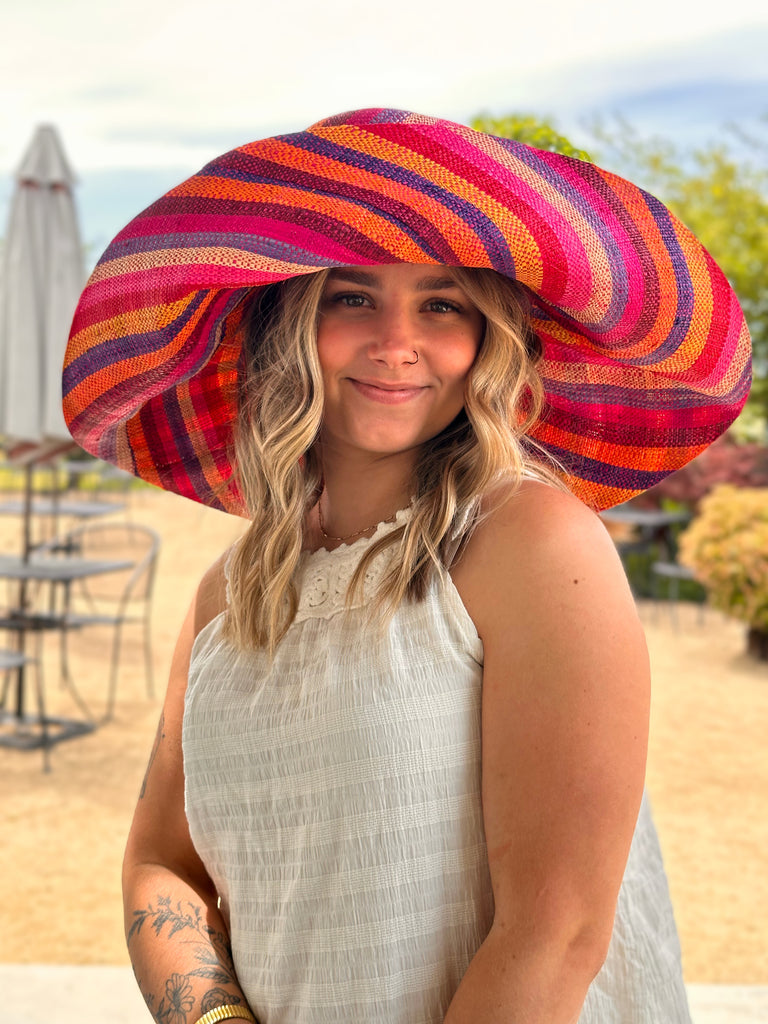 Model wearing 5" & 7" Wide Fiesta Stripe Multicolor Stripes Packable Straw Sun Hat Handmade loomed raffia multi width bands of fuchsia pink, bordeaux red, saffron yellow, coral orange, natural straw color, purple, blue, etc. create a swirl pattern - Shebobo