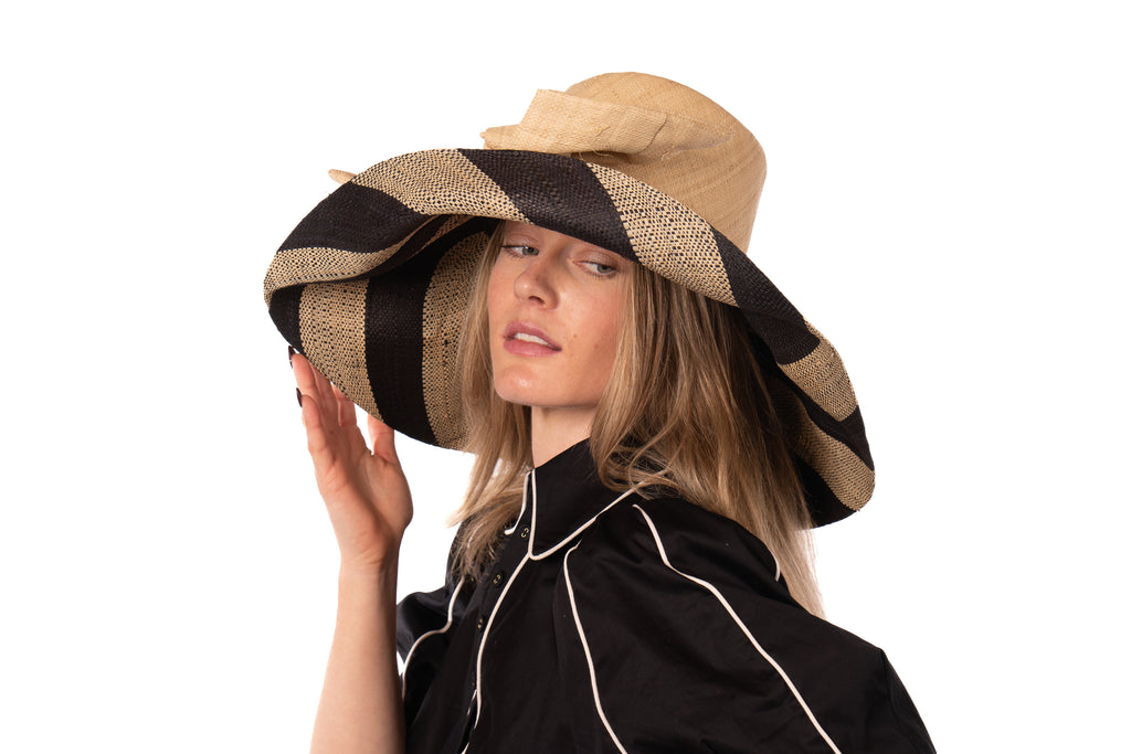 Model wearing 7" Wide Brim Natural Two Tone Wide Stripe Straw Sun Hats with Big Bow handmade loomed raffia palm fiber in a solid hue of Natural on the crown and matching oversized big bow embellishment hat band, with two tone even width wide stripe pattern of black and natural straw color that creates a swirl patterned brim - Shebobo