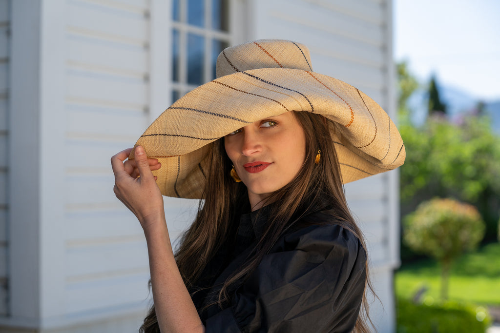 Model wearing 7 inch wide brim natural and Neutrals multicolor pinstripes packable raffia straw hat handmade loomed raffia in wide bands of natural straw color with narrow bands of blush orange/pink, cinnamon/tobacco/brown, black, and grey create a stripe swirl pattern - Shebobo