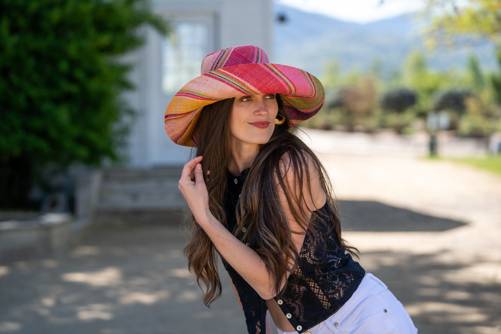 Model wearing 5" Wide Brim Raspberry Swirl Multicolor Stripes Packable Straw Sun Hat Handmade loomed raffia in multi width bands of purple, light pink, coral orange/red, lime green, natural straw color, light blue, yellow, etc. create a swirl pattern - Shebobo
