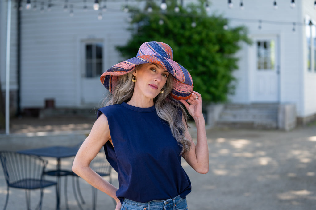 Model wearing 7" Wide Brim Retro Swirl Packable Straw Sun Hat handmade loomed raffia in multi width stripes of denim retro/light/blue, coral orange/red, dusk navy/dark/blue, and light pink create a swirl pattern - Shebobo