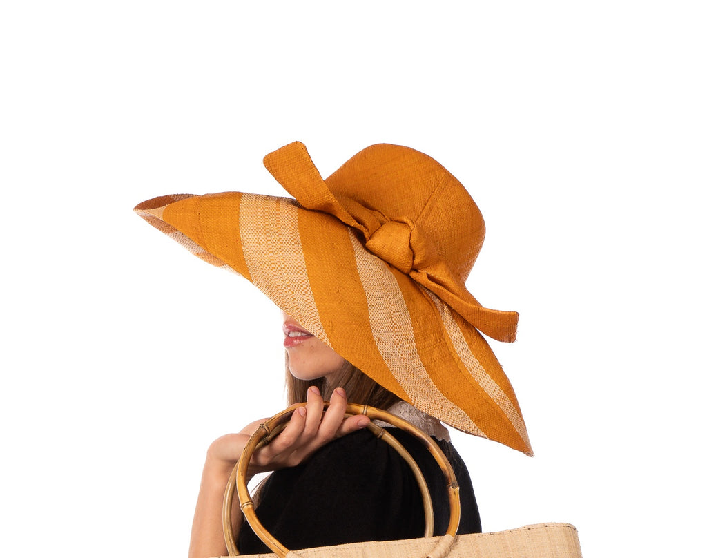 Model wearing 7" Wide Brim Saffron Two Tone Wide Stripe Straw Sun Hats with Big Bow handmade loomed raffia palm fiber in a solid hue of Saffron yellow/orange on the crown and matching oversized big bow embellishment hat band, with two tone even width wide stripe pattern of saffron and natural straw color that creates a swirl patterned brim - Shebobo