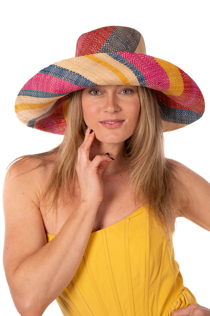 Model wearing 7" Wide Brim Sassafras Swirl Multicolor Stripes Packable Straw Sun Hat Handmade loomed raffia in multi width bands of fuchsia pink, red, saffron yellow, navy blue, and natural straw color create a swirl pattern - Shebobo