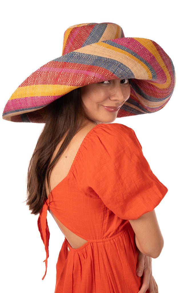 Model wearing 7" Wide Brim Sassafras Swirl Multicolor Stripes Packable Straw Sun Hat Handmade loomed raffia in multi width bands of fuchsia pink, red, saffron yellow, navy blue, and natural straw color create a swirl pattern - Shebobo