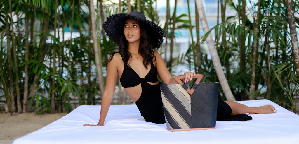 Model wearing 5 inch wide brim handmade woven raffia palm black straw sun hat with raw fringe edge and Zuki Two Tone Multicolor Straw Handbag in Black Swirl multi width diagonal stripe of black, grey, and natural straw color on one vertical half of the bag and solid black on the other half with a carved wooden triangle handle - Shebobo