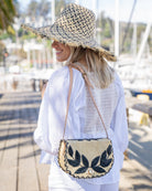 Model wearing 70's Crossbody Straw Bag handmade loomed natural raffia palm fiber purse with black accent cross stitch edging and matching leaf design embroidery handbag with adjustable leather strap - Shebobo (with 5" Brim Kat Black and Natural Multicolor Straw Hat)