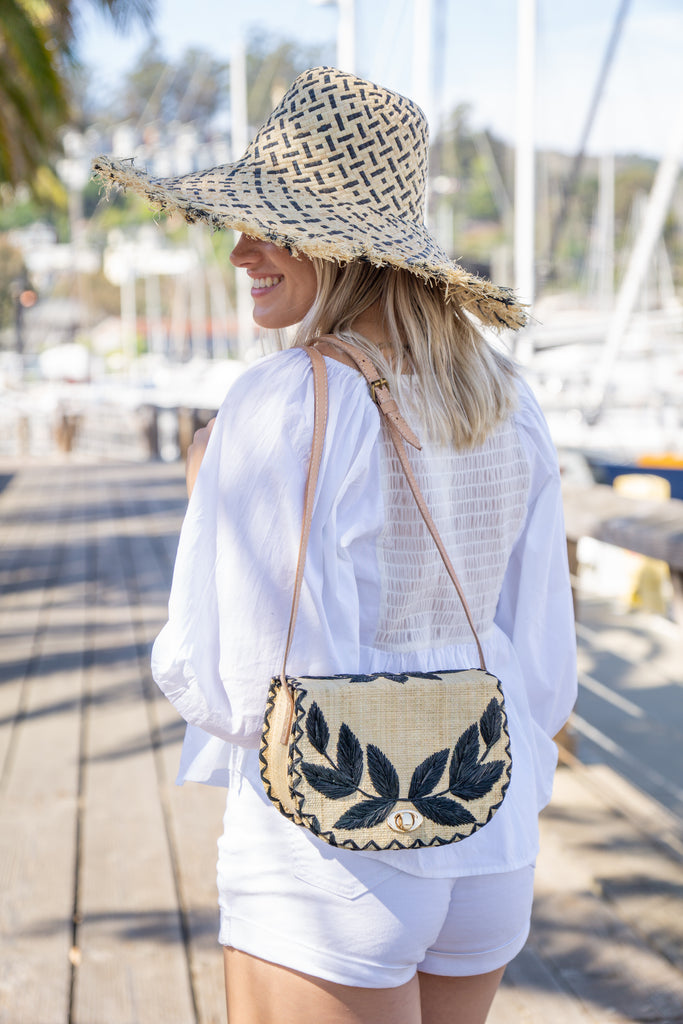 Model wearing 70's Crossbody Straw Bag handmade loomed natural raffia palm fiber purse with black accent cross stitch edging and matching leaf design embroidery handbag with adjustable leather strap - Shebobo (with 5" Brim Kat Black and Natural Multicolor Straw Hat)