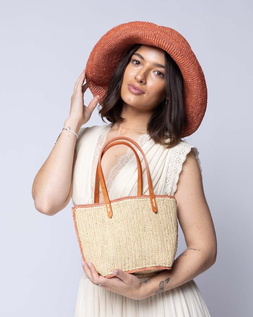 Model wearing Side view Adele Mini Crochet Raffia Straw Handbag Purse with Colored Edge Natural & Blush handmade crochet raffia palm fiber in a solid hue of natural straw color with contrasting narrow band of blush orange/pink around the outer edges of the bag with subtle vertical band texture and leather handles - Shebobo