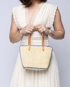 Model wearing Adele Mini Crochet Raffia Straw Handbag Purse with Colored Edge Natural & Denim handmade crochet raffia palm fiber in a solid hue of natural straw color with contrasting narrow band of denim light/blue around the outer edges of the bag with subtle vertical band texture and leather handles - Shebobo