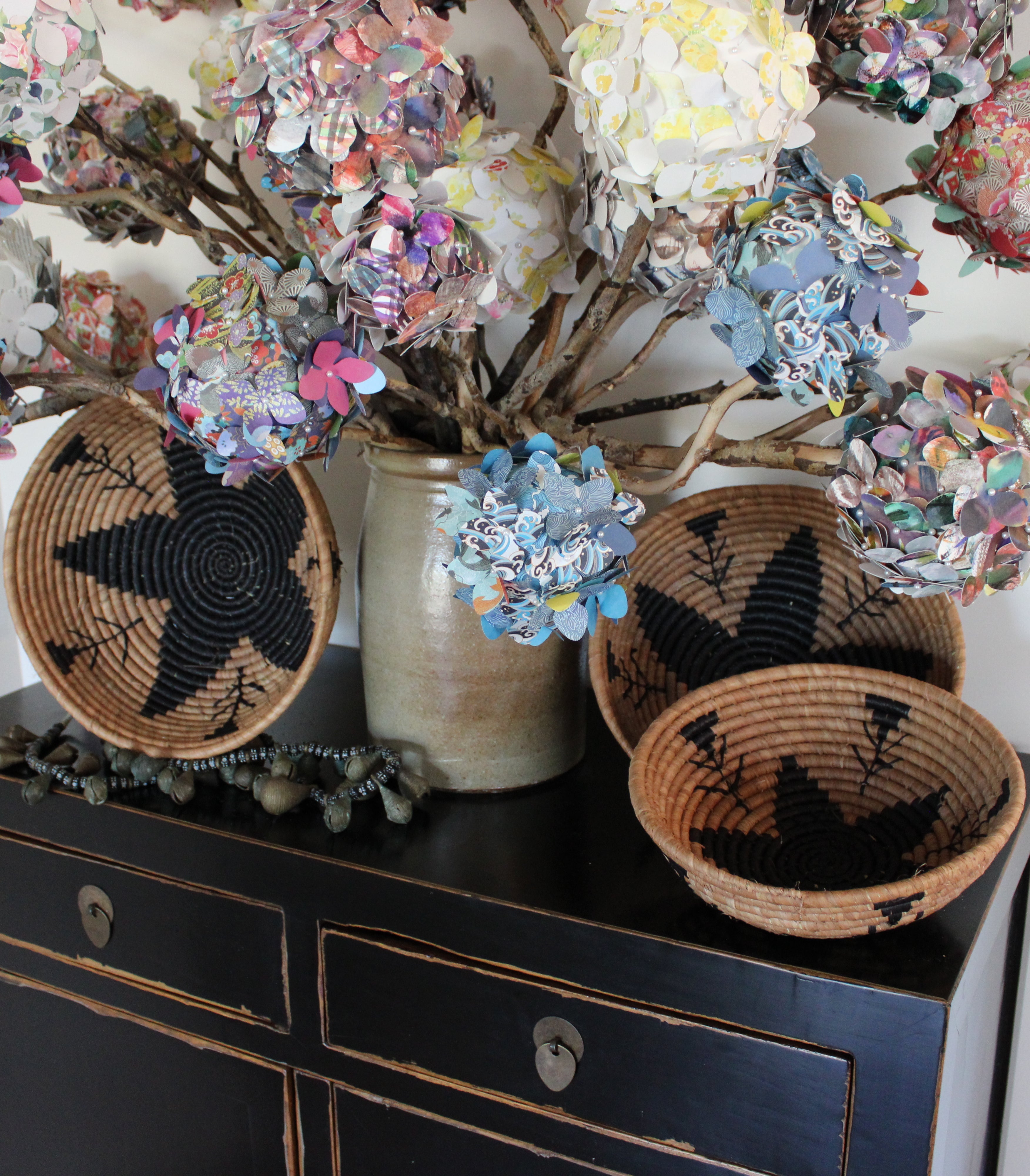 Home decor lifestyle view Amara Raffia Straw Decor Display Basket Bowls: Set of 3 handmade woven natural raffia palm fiber in a crochet loop technique concentric coiled circular pattern with centered black star or flower in the bottom of the bowl that goes up the cinnamon/tobacco/brown sides with matching black flower/arrow pattern on sides set of small, medium, and large straw display bowls - Shebobo