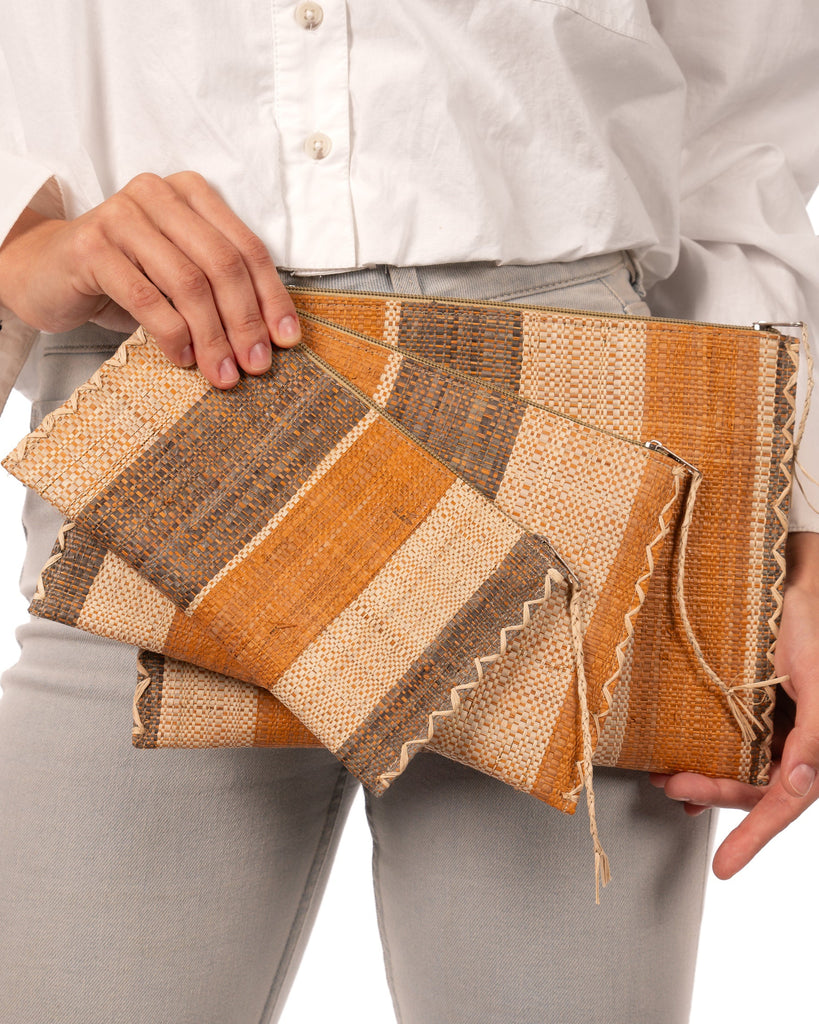 Model wearing Amelia Set of 3 Nesting Zippered Straw Clutches Blush Swirl Multicolor Stripe Pattern handmade loomed raffia in multiple widths of vertical stripes in natural, blush orange/pink, and grey with matching zipper and braided zipper pull with cross stitch edging in three sizes of small, medium, and large - Shebobo