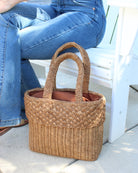 Model wearing Anna Petal Pattern Crochet Straw Basket Handbag handmade natural raffia palm fiber in a solid hue of Cappuccino tea/light/brown crochet in multiple textures resulting in a subtle pattern of vertical linked bands on the bottom 2/3 of the bag, and a floral petal or shell pattern on the upper third with rolled panel of links making up the handles Beach Bag Purse - Shebobo