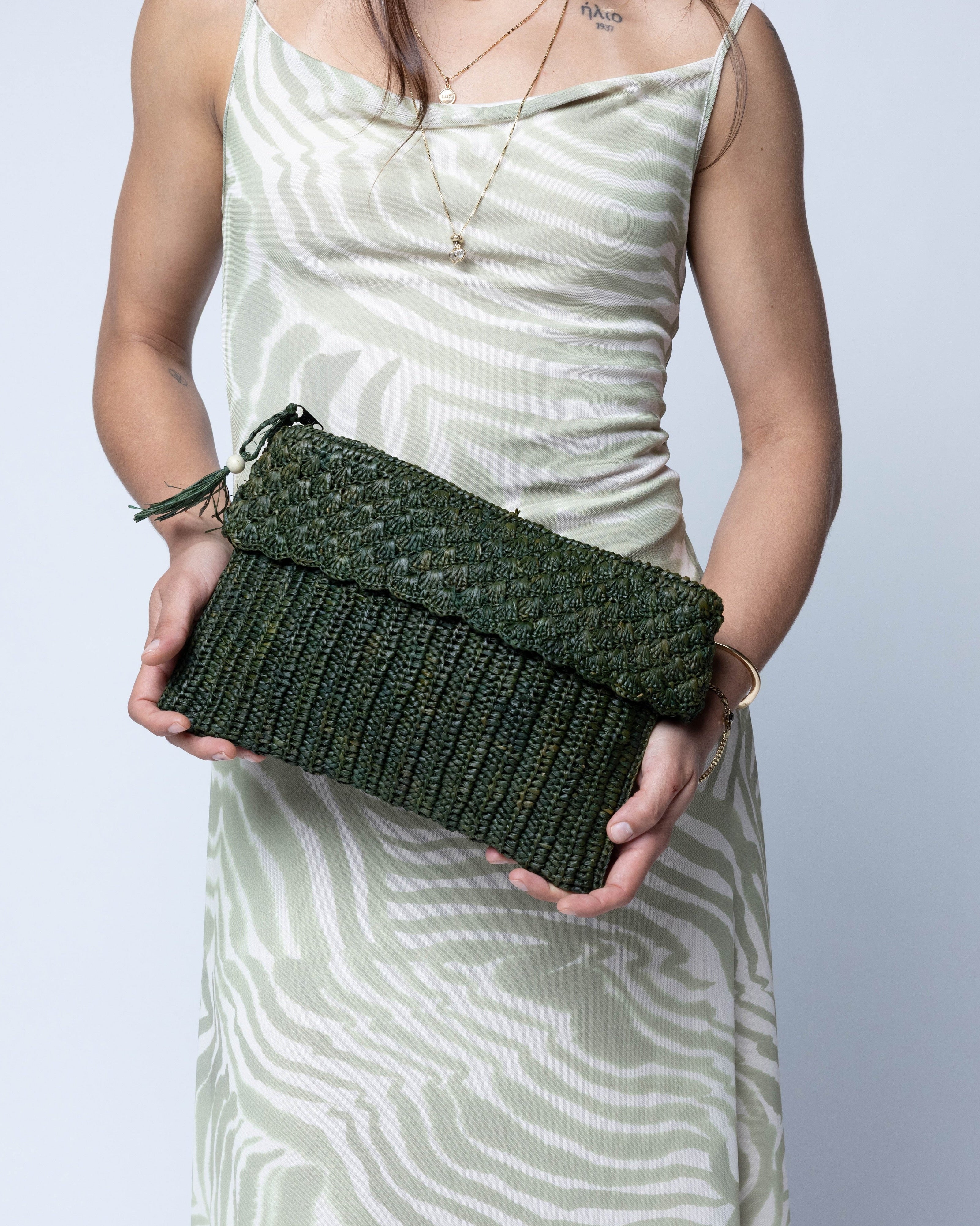Model wearing Anna Petal Pattern Crochet Straw Pouch Clutch Purse handmade natural raffia palm fiber in a solid hue of Deep Green dark/forrest/green crochet in multiple textures resulting in a subtle pattern of vertical linked bands on the bottom half of the bag, and a floral petal or shell pattern on the upper half - Shebobo