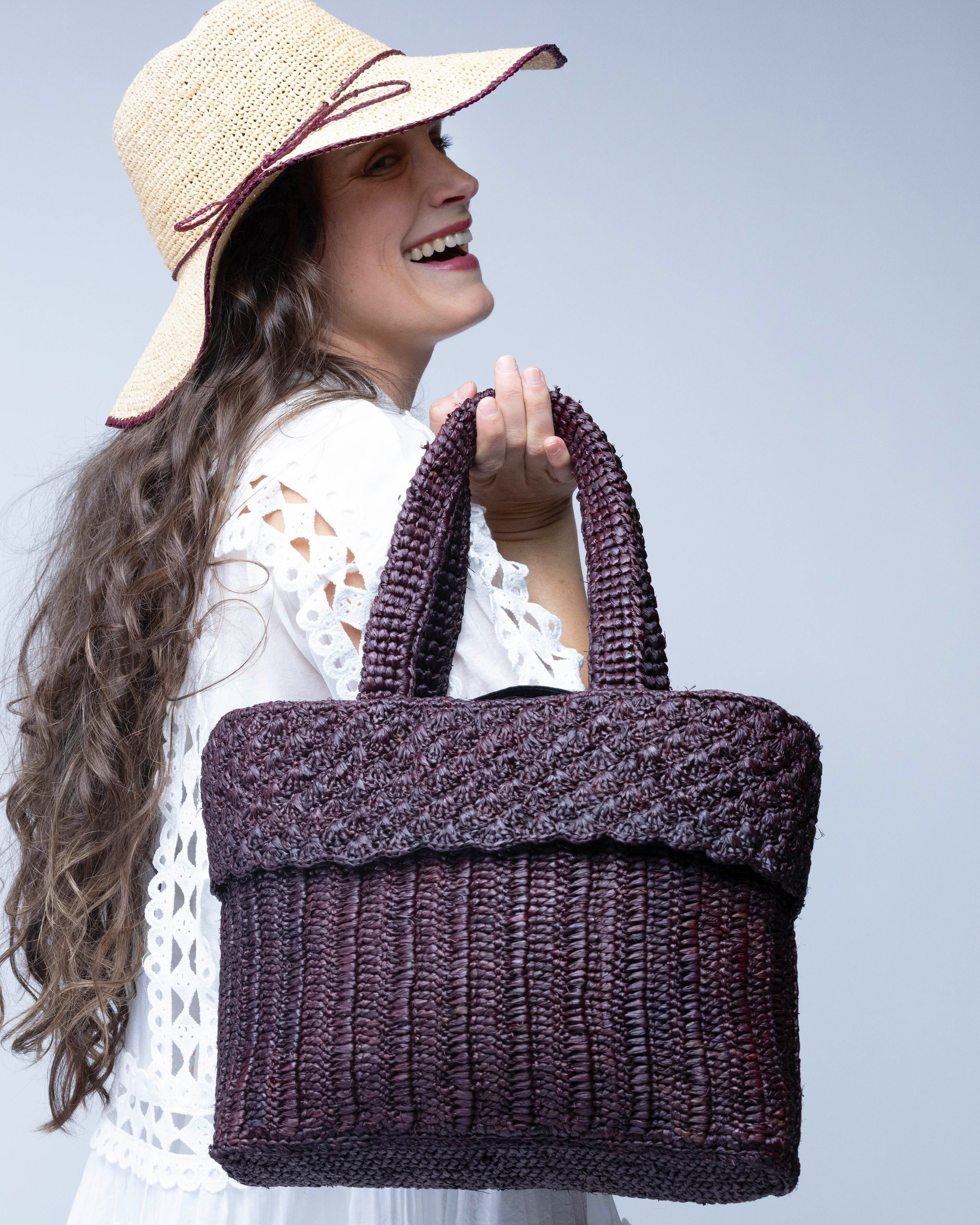 Model wearing Anna Petal Pattern Crochet Straw Basket Handbag handmade natural raffia palm fiber in a solid hue of Espresso chocolate/dark/brown crochet in multiple textures resulting in a subtle pattern of vertical linked bands on the bottom 2/3 of the bag, and a floral petal or shell pattern on the upper third with rolled panel of links making up the handles Beach Bag Purse - Shebobo (with Rachel 4" Brim Crochet Straw Sun Hats with Colored Edge)