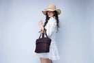 Model wearing Anna Petal Pattern Crochet Straw Basket Handbag handmade natural raffia palm fiber in a solid hue of Mocha chocolate/dark/brown crochet in multiple textures resulting in a subtle pattern of vertical linked bands on the bottom 2/3 of the bag, and a floral petal or shell pattern on the upper third with rolled panel of links making up the handles Beach Bag Purse - Shebobo (with Rachel 4" Brim Crochet Straw Sun Hats with Colored Edge)