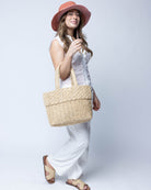 Model wearing Anna Petal Pattern Crochet Straw Basket Handbag handmade natural raffia palm fiber in a solid hue of Natural straw color crochet in multiple textures resulting in a subtle pattern of vertical linked bands on the bottom 2/3 of the bag, and a floral petal or shell pattern on the upper third with rolled panel of links making up the handles Beach Bag Purse - Shebobo (with Sequoia 3" Brim Swirl Crochet Panama Unisex Straw Hats and Lizzie Straw Sandal Shoes)