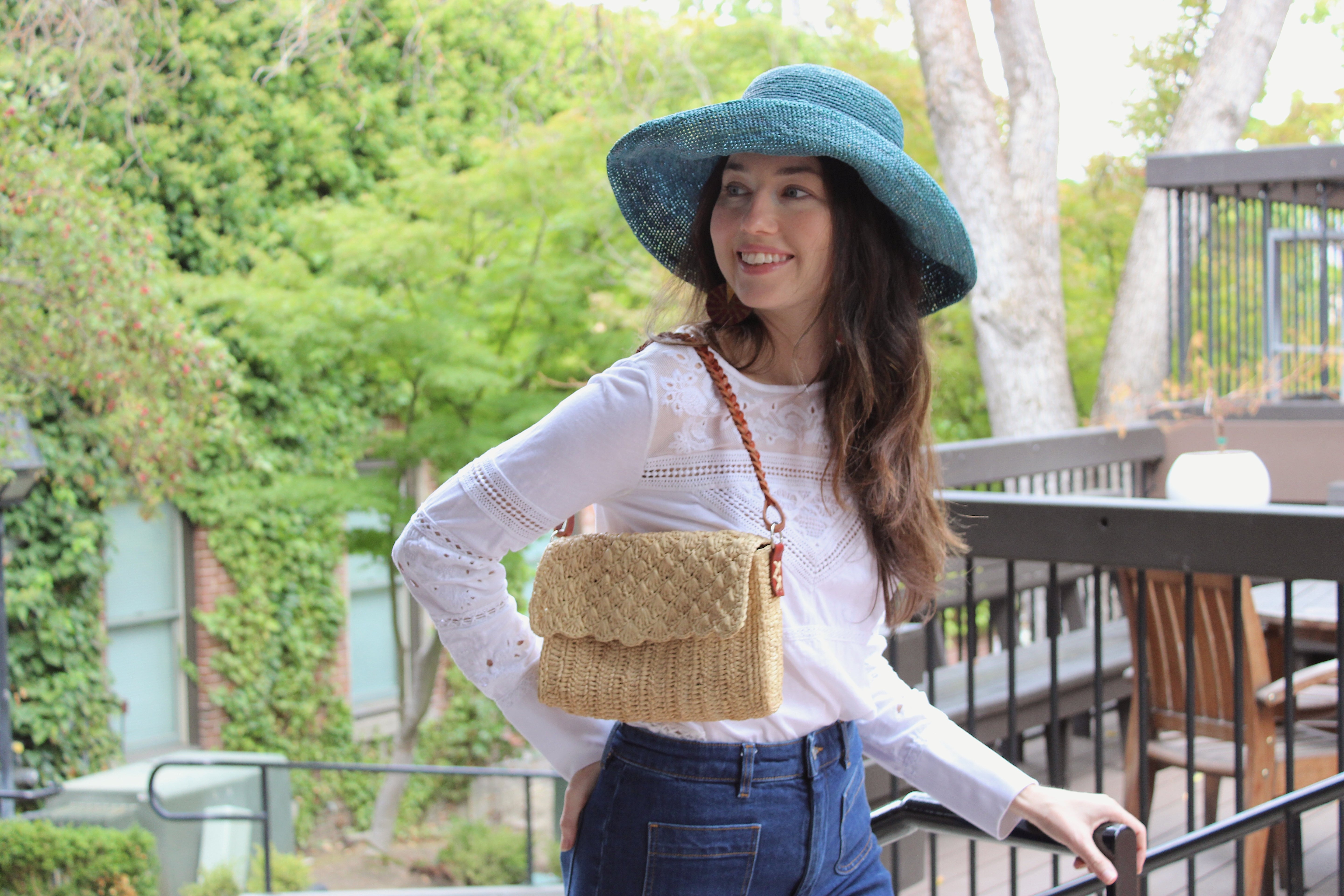 Model wearing Anna Petal Pattern Crochet Straw Over Shoulder Envelope Clutch Purse handmade raffia palm fiber in a solid hue of Natural straw color crochet in multiple textures resulting in a subtle pattern of vertical linked bands on the bottom half of the bag, and a floral petal or shell pattern on the upper envelope flap with braided leather shoulder strap handbag shoulder bag - Shebobo (with Leor 3" Brim Crochet Straw Sun Hats)
