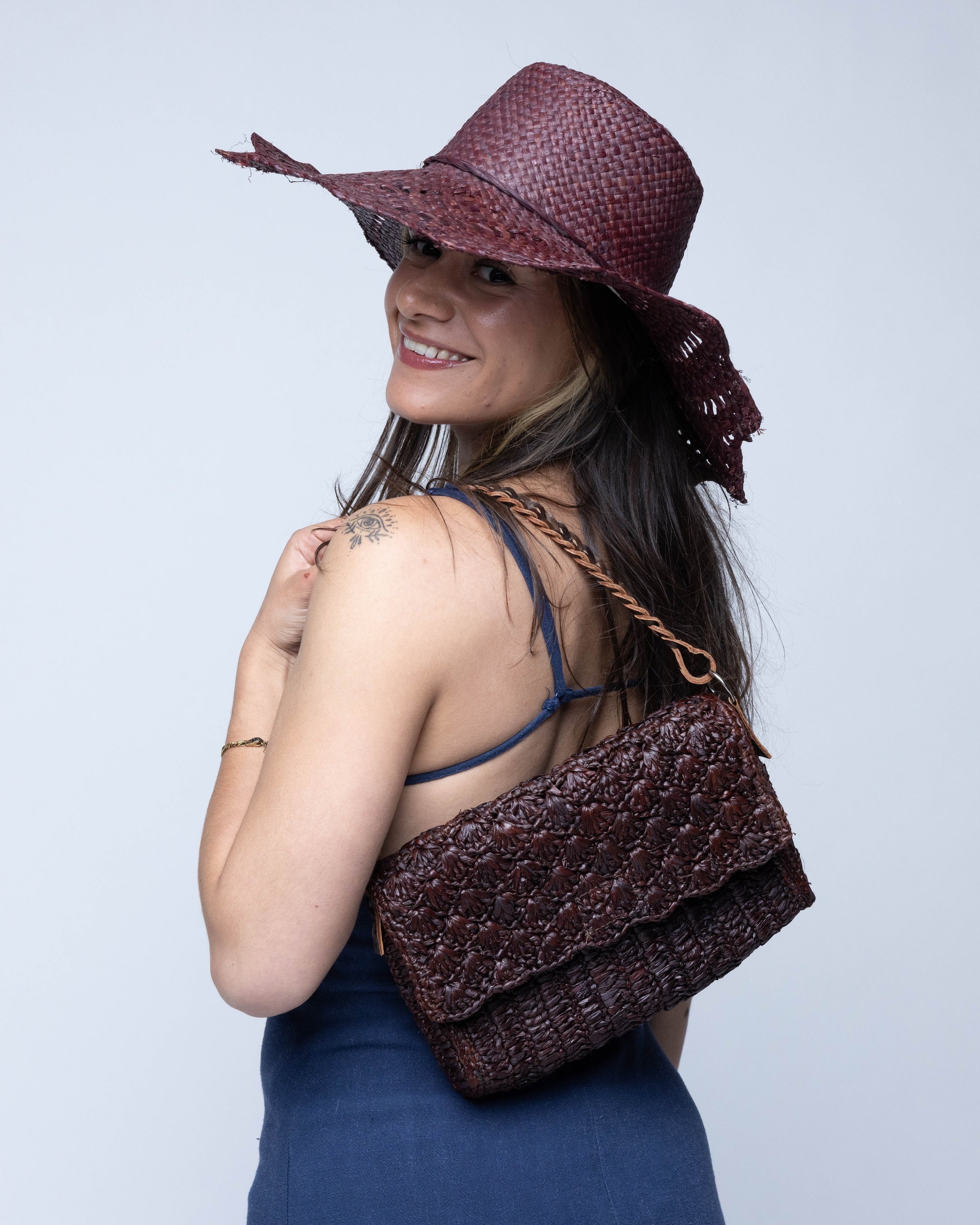 Model wearing Anna Petal Pattern Crochet Straw Over Shoulder Envelope Clutch Purse handmade raffia palm fiber in a solid hue of Mocha chocolate/dark/brown crochet in multiple textures resulting in a subtle pattern of vertical linked bands on the bottom half of the bag, and a floral petal or shell pattern on the upper envelope flap with braided leather shoulder strap handbag shoulder bag - Shebobo (with Sara 4.5" Brim Woven Straw Sun Hats with Zigzag Edge)