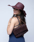 Model wearing Anna Petal Pattern Crochet Straw Over Shoulder Envelope Clutch Purse handmade raffia palm fiber in a solid hue of Mocha chocolate/dark/brown crochet in multiple textures resulting in a subtle pattern of vertical linked bands on the bottom half of the bag, and a floral petal or shell pattern on the upper envelope flap with braided leather shoulder strap handbag shoulder bag - Shebobo (with Sara 4.5" Brim Woven Straw Sun Hats with Zigzag Edge)