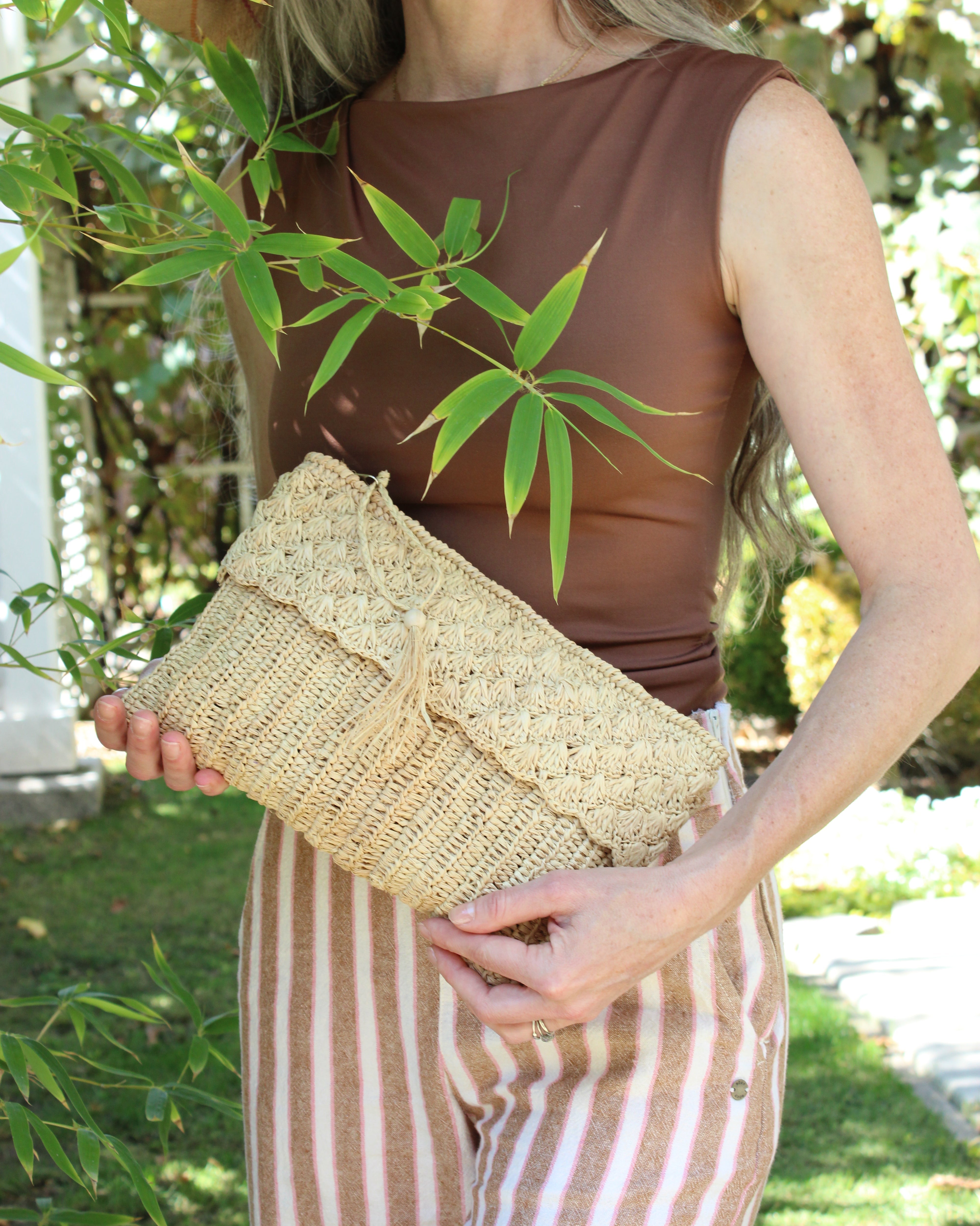 Model wearing Anna Petal Pattern Crochet Straw Pouch Clutch Purse handmade natural raffia palm fiber in a solid hue of Natural straw color crochet in multiple textures resulting in a subtle pattern of vertical linked bands on the bottom half of the bag, and a floral petal or shell pattern on the upper half - Shebobo
