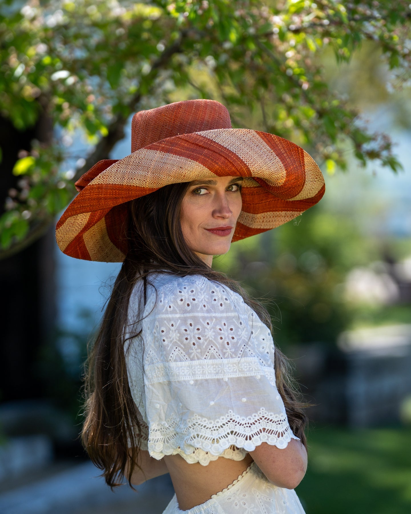 Model wearing Audrey 7" Wide Brim Whisky Two Tone Wide Stripe Straw Sun Hats with Big Bow handmade loomed raffia palm fiber in a solid hue of whisky carmel/orange/red/brown on the crown and matching oversized big bow embellishment hat band, with two tone even width wide stripe pattern of coral and natural straw color that creates a swirl patterned brim - Shebobo