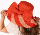 Model wearing Audrey 5" Wide Brim Coral Two Tone Wide Stripe Straw Sun Hats with Big Bow handmade loomed raffia palm fiber in a solid hue of Coral orange/red on the crown and matching oversized big bow embellishment hat band, with two tone even width wide stripe pattern of coral and natural straw color that creates a swirl patterned brim - Shebobo