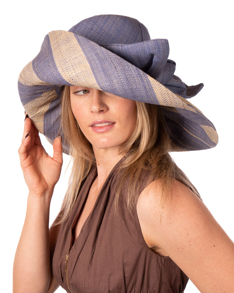 Model wearing Audrey 7" Wide Brim Denim Two Tone Wide Stripe Straw Sun Hats with Big Bow handmade loomed raffia palm fiber in a solid hue of Denim retro/light/blue on the crown and matching oversized big bow embellishment hat band, with two tone even width wide stripe pattern of denim and natural straw color that creates a swirl patterned brim - Shebobo
