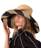 Model wearing Audrey 7" Wide Brim Natural Two Tone Wide Stripe Straw Sun Hats with Big Bow handmade loomed raffia palm fiber in a solid hue of Natural on the crown and matching oversized big bow embellishment hat band, with two tone even width wide stripe pattern of black and natural straw color that creates a swirl patterned brim - Shebobo