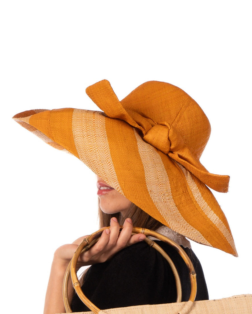 Model wearing Audrey 7" Wide Brim Saffron Two Tone Wide Stripe Straw Sun Hats with Big Bow handmade loomed raffia palm fiber in a solid hue of Saffron yellow/orange on the crown and matching oversized big bow embellishment hat band, with two tone even width wide stripe pattern of saffron and natural straw color that creates a swirl patterned brim - Shebobo