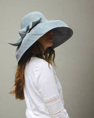 Model wearing Audrey 7" Wide Brim Solid Color Straw Sun Hats with Big Bow handmade loomed natural raffia straw fiber in a solid hue of Denim retro/light/blue with matching oversized big bow embellishment hat band - Shebobo