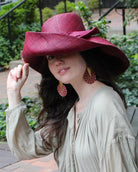 Model wearing Audrey 7" Wide Brim Solid Color Straw Sun Hats with Big Bow handmade loomed natural raffia straw fiber in a solid hue of Umberwine bordeaux/claret/deep/dark/red/purple with matching oversized big bow embellishment hat band - Shebobo