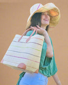 Model wearing Augustine pinstripes straw basket bag handmade loomed raffia in natural wide horizontal bands with multicolor bright colors of coral red/orange, bordeaux dark red, lime, grey, and fuchsia pink small bands running through in a stripe pattern with leather accents and handles handbag purse with Monica 7" Packable Brights Multicolor Pinstripe Straw Sun Hat - Shebobo