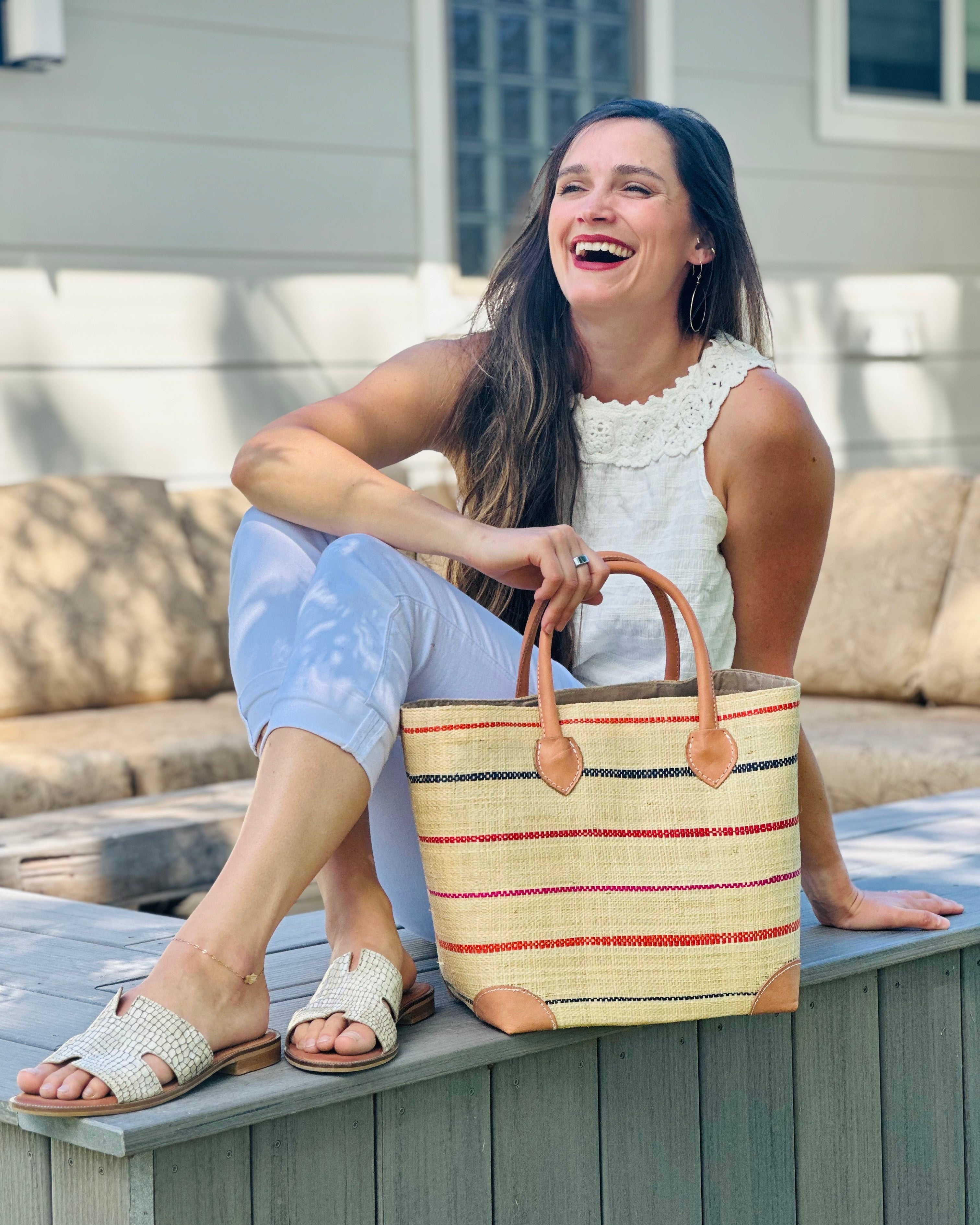 Model wearing Augustine pinstripes straw basket bag handmade loomed raffia in natural wide horizontal bands with multicolor coral red/orange, fuchsia pink, and navy blue small bands running through in a stripe pattern with leather accents and handles handbag purse in size medium - Shebobo