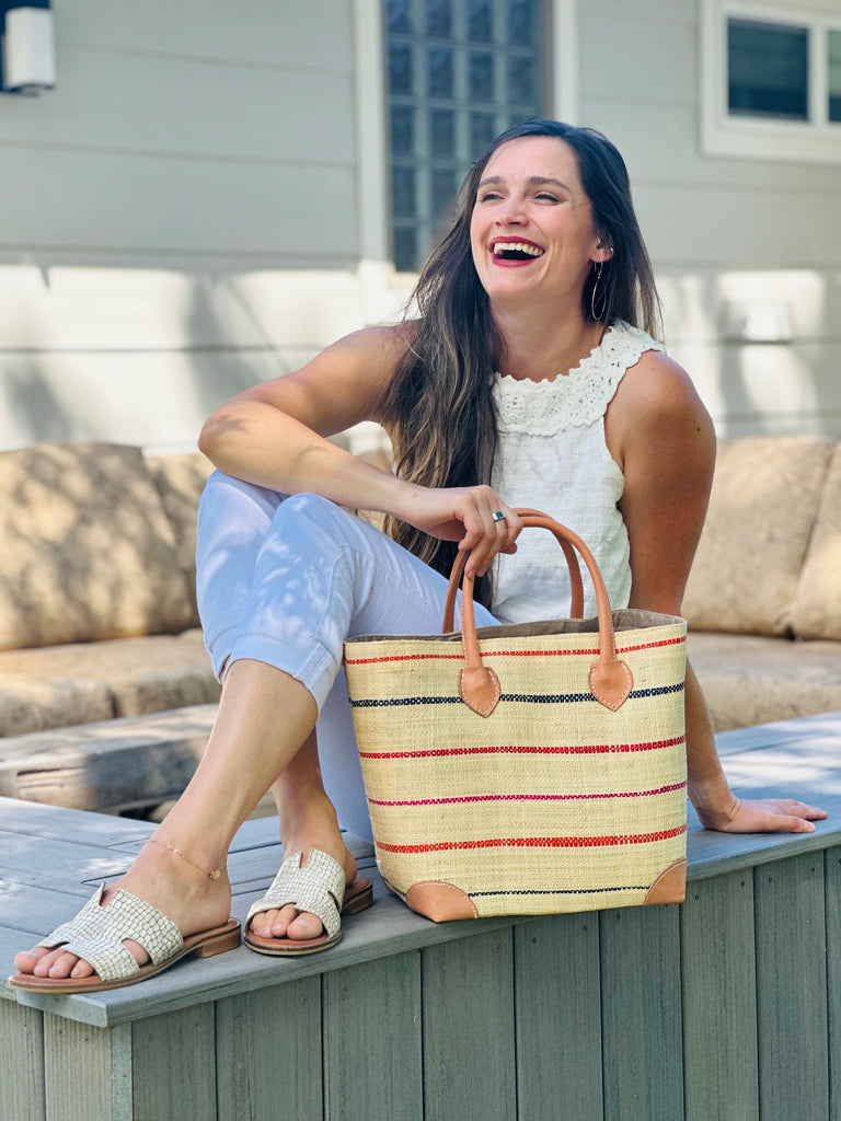 Model wearing Augustine pinstripes straw basket bag handmade loomed raffia in natural wide horizontal bands with multicolor coral red/orange, fuchsia pink, and navy blue small bands running through in a stripe pattern with leather accents and handles handbag purse in size medium - Shebobo