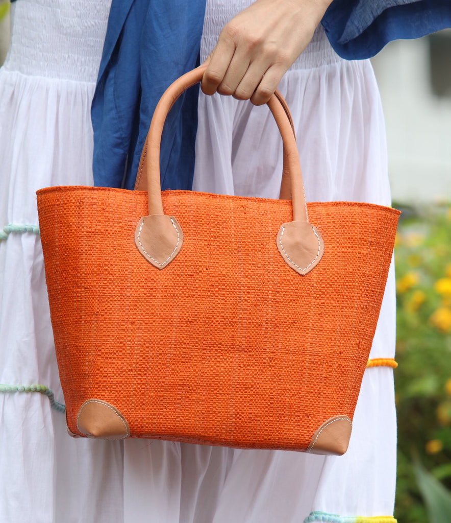 Model wearing Augustine Straw Basket Bag handmade loomed raffia handbag with leather corners and handles in coral orange/red - Shebobo