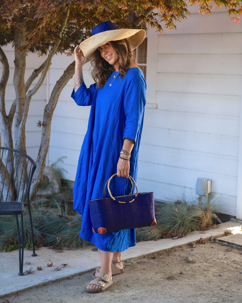 Model wearing Bebe Straw Handbag with Bamboo Handle handmade loomed natural raffia palm fiber in a solid hue of Dusk dark/navy/blue with molded circular bamboo handles, leather feet, and assorted print African wax-cloth fabric liner - Shebobo (with Monica 5" & 7" Wide Brim Two Tone Packable Straw Sun Hats)