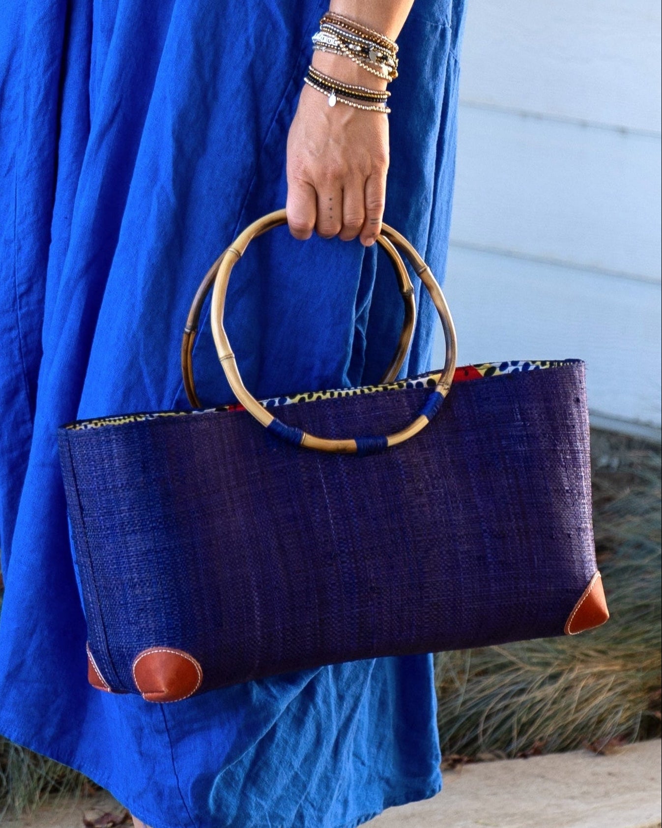Model wearing Bebe Straw Handbag with Bamboo Handle handmade loomed natural raffia palm fiber in a solid hue of Dusk dark/navy/blue with molded circular bamboo handles, leather feet, and assorted print African wax-cloth fabric liner - Shebobo