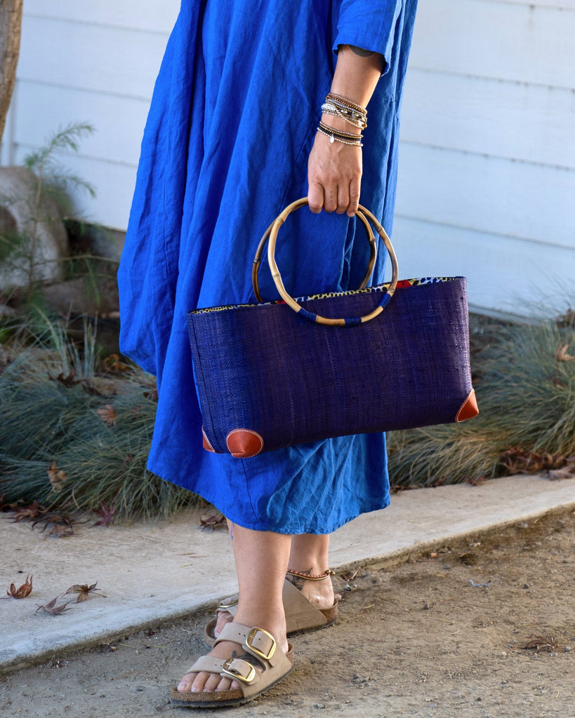 Model wearing Bebe Straw Handbag with Bamboo Handle handmade loomed natural raffia palm fiber in a solid hue of Dusk dark/navy/blue with molded circular bamboo handles, leather feet, and assorted print African wax-cloth fabric liner - Shebobo