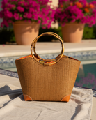 Lifestyle view Betty Straw Bucket Bag with Bamboo Handles handmade loomed natural raffia palm fiber in a solid hue of tobacco cinnamon/dark/brown with circular bamboo handles, leather feet, and colorful African Print wax cloth liner handbag purse tote - Shebobo