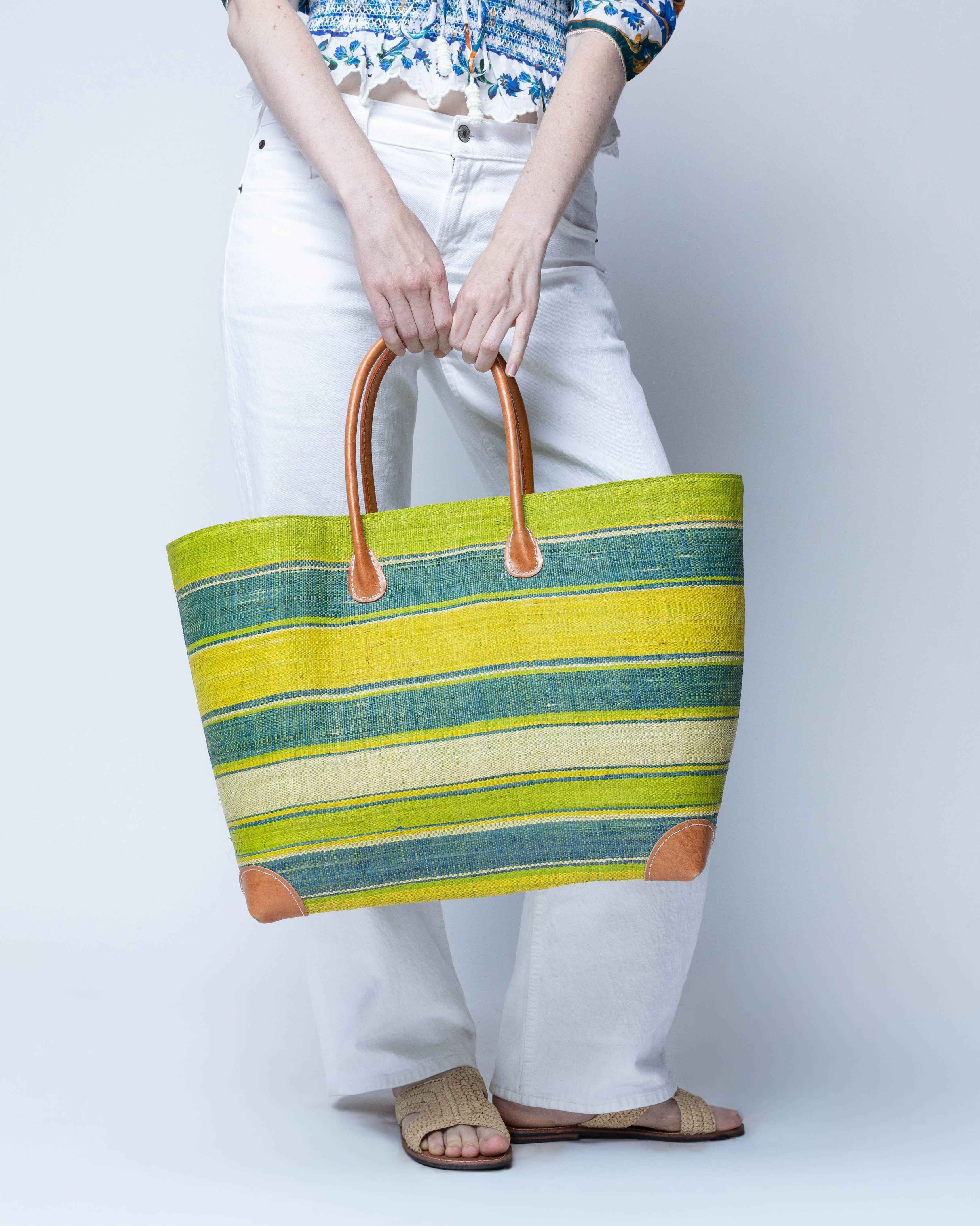 Model wearing Bolinas Izarra Swirl XL Straw Basket Bag handmade loomed natural raffia palm fiber in a multicolor horizontal stripe pattern of multiple width bands of natural straw color, lime green, turquoise blue, navy blue, yellow, etc. with leather handles and accents large tote bag shoulder bag handbag beach bag - Shebobo