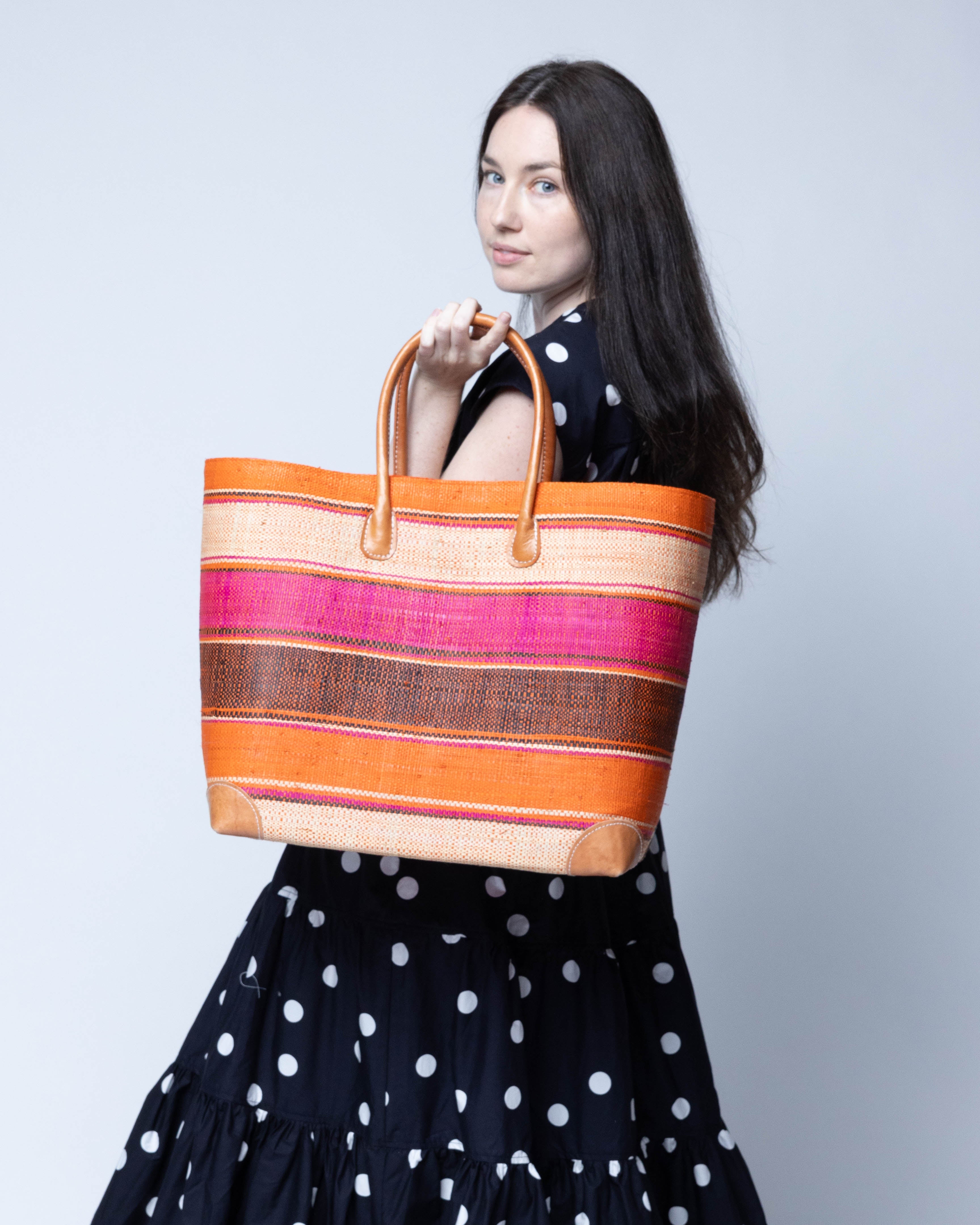 Model wearing Bolinas Whimsy Swirl XL Straw Basket Bag handmade loomed natural raffia palm fiber in a multicolor horizontal stripe pattern of multiple width bands of natural straw color, fuchsia hot/bright/barbie/pink, coral orange/red, black, etc. with leather handles and accents large tote bag shoulder bag handbag beach bag - Shebobo