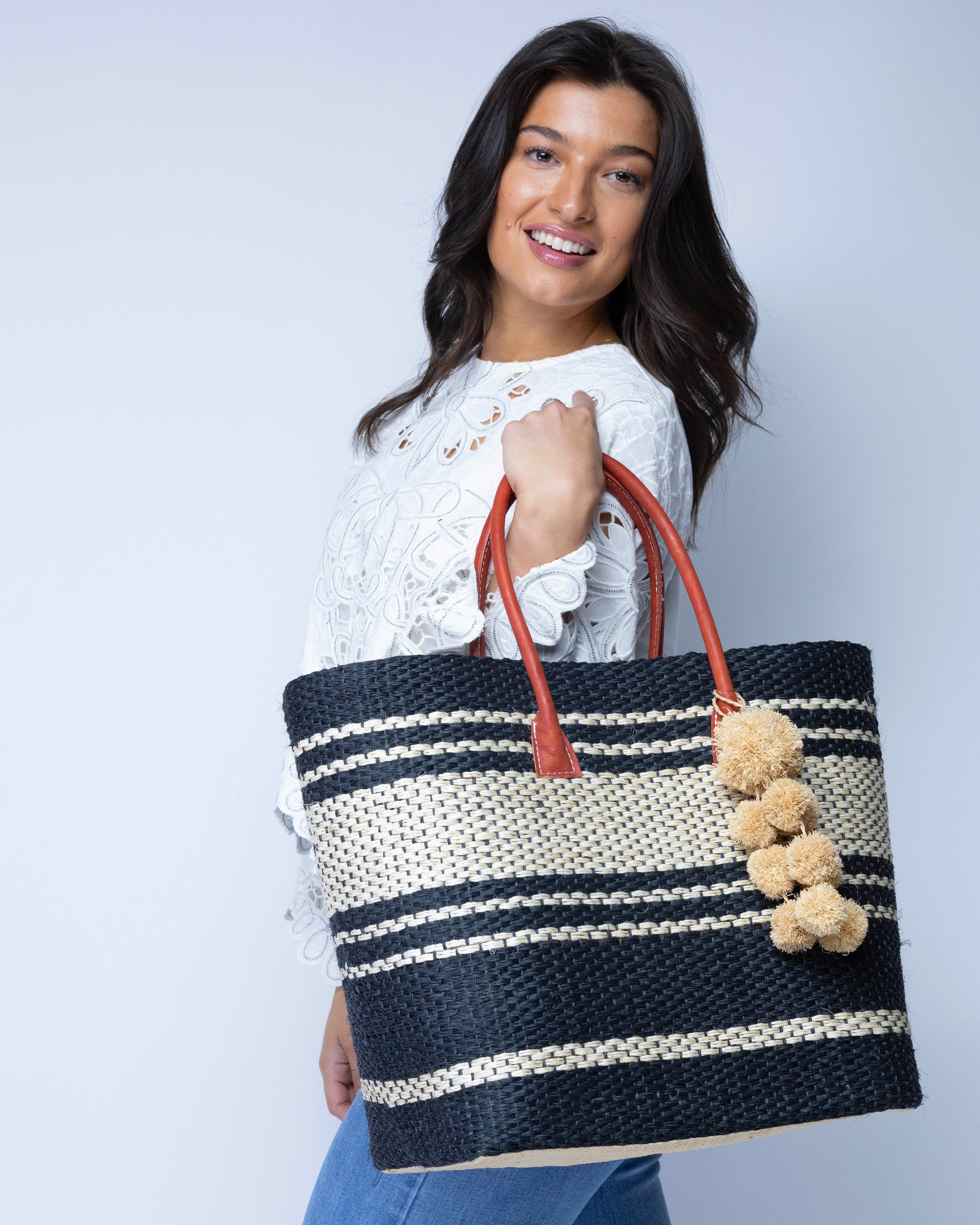 Model wearing Cabrillo Sisal Basket Bag with Waterfall Pompoms Charm Embellishment handmade natural sisal fiber woven into a multicolor two tone Black and natural straw color  horizontal stripe pattern with muti-width bands of color plus leather handles and natural raffia charm shoulder bag beach bag - Shebobo