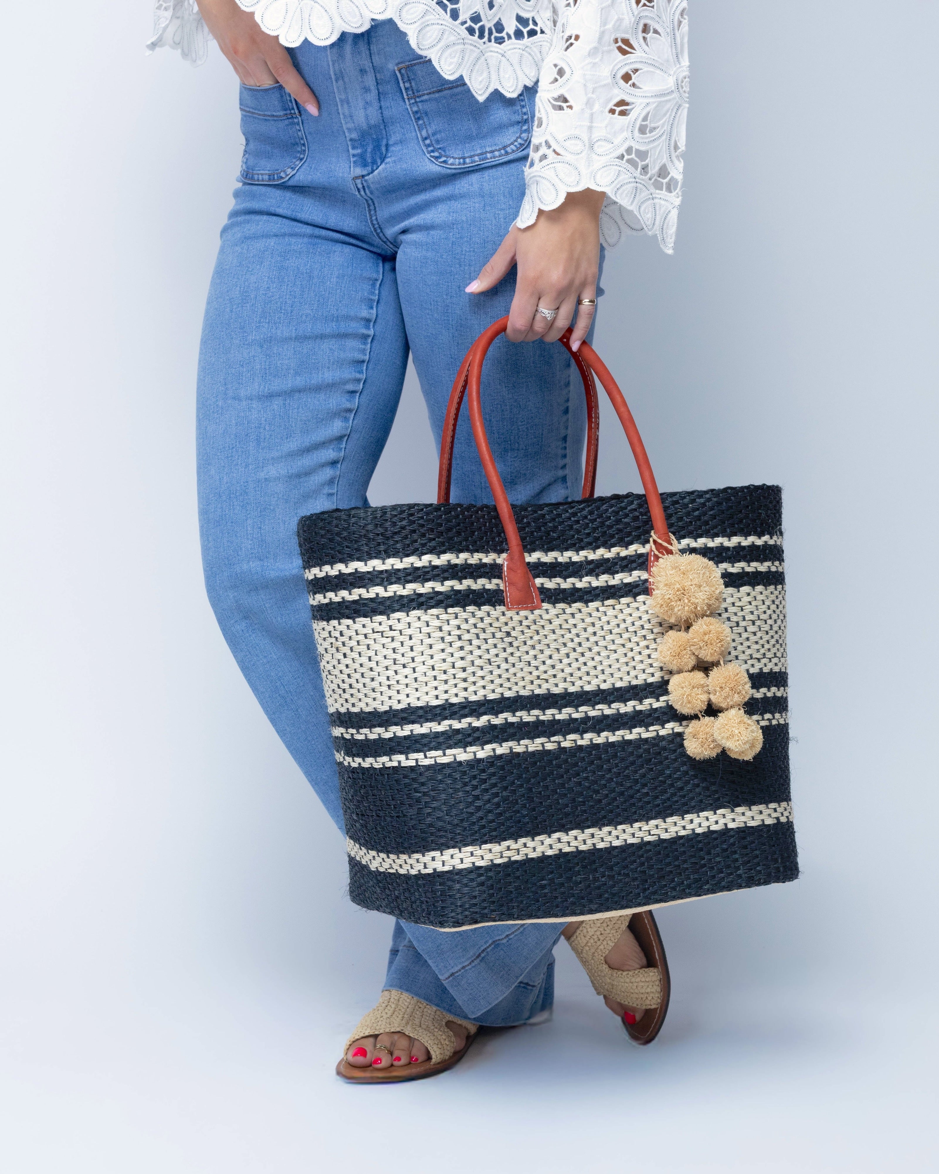 Model wearing Cabrillo Sisal Basket Bag with Waterfall Pompoms Charm Embellishment handmade natural sisal fiber woven into a multicolor two tone Black and natural straw color  horizontal stripe pattern with muti-width bands of color plus leather handles and natural raffia charm shoulder bag beach bag - Shebobo