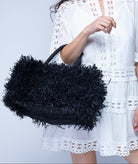 Model wearing Calypso Pompom Pouf Crochet Straw Basket Handbag handmade natural raffia palm fiber in a solid hue of Black with brushed fringe strands crochet and knotted into a pouf texture with matching wrapped handles purse - Shebobo