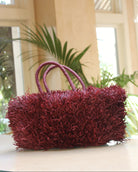 Lifestyle view Calypso Pompom Pouf Crochet Straw Basket Handbag handmade natural raffia palm fiber in a solid hue of Umberwine burgunday/bordeaux/dark/purple/red with brushed fringe strands crochet and knotted into a pouf texture with matching wrapped handles purse - Shebobo