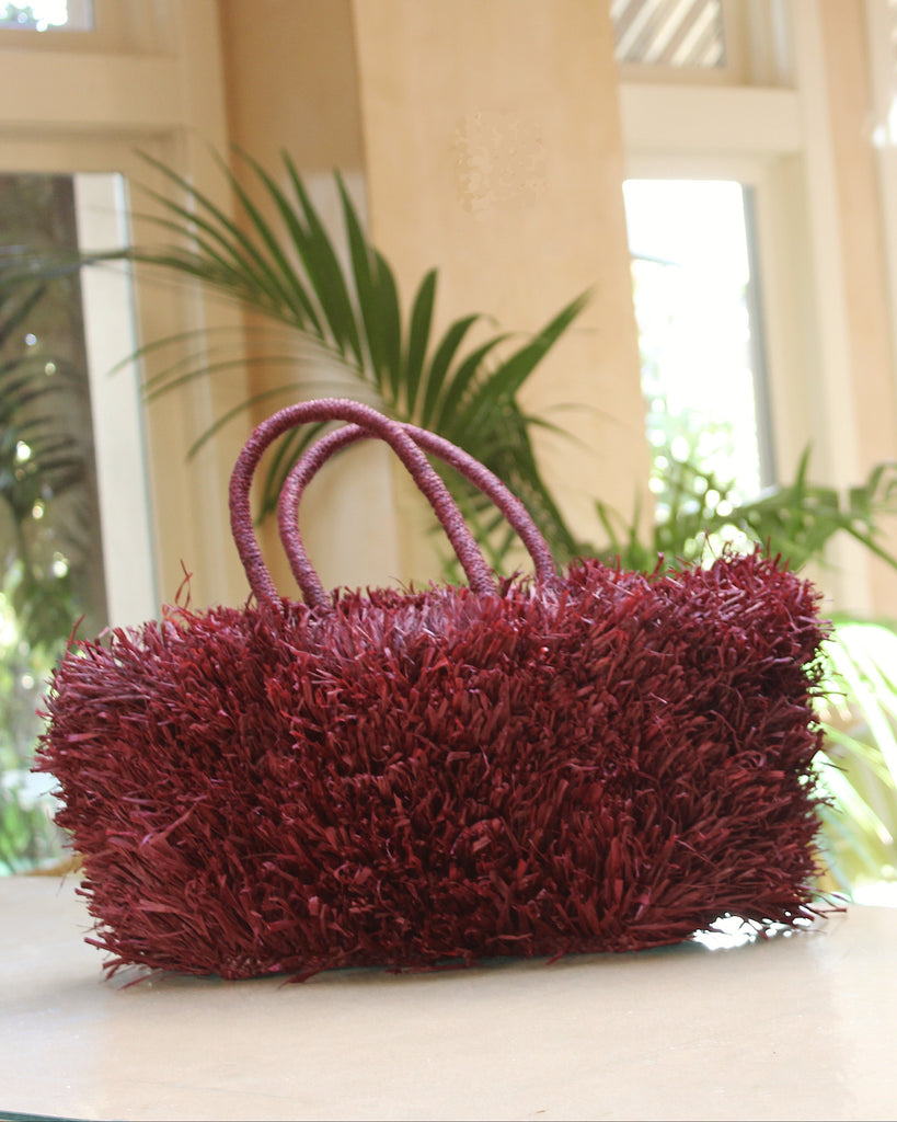 Lifestyle view Calypso Pompom Pouf Crochet Straw Basket Handbag handmade natural raffia palm fiber in a solid hue of Umberwine burgunday/bordeaux/dark/purple/red with brushed fringe strands crochet and knotted into a pouf texture with matching wrapped handles purse - Shebobo